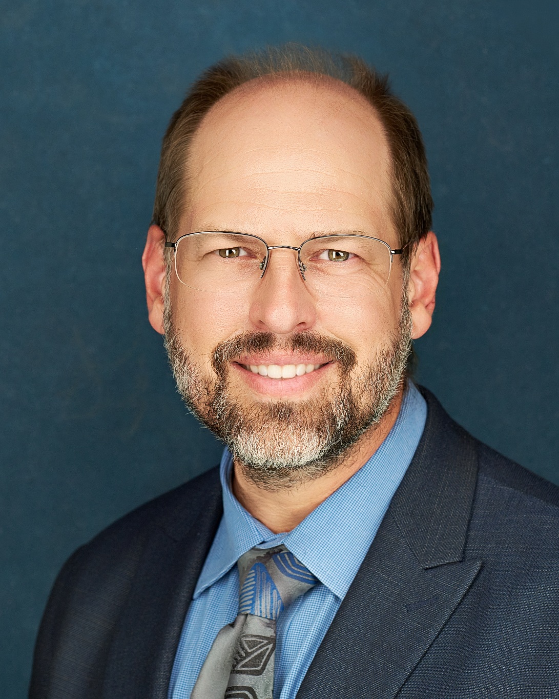 Professional headshot of Ken Brey wearing a suit and tie, smiling at the camera.